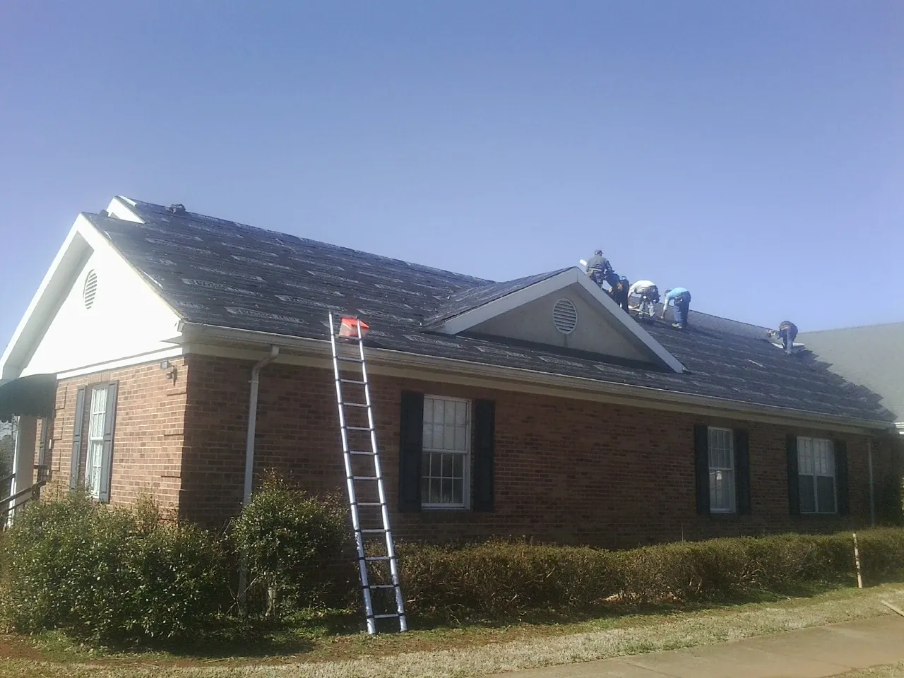 Crew tearing shingles off a brick ranch roof with a ladder set — Emergency Roof Repair work by our crew in Locust