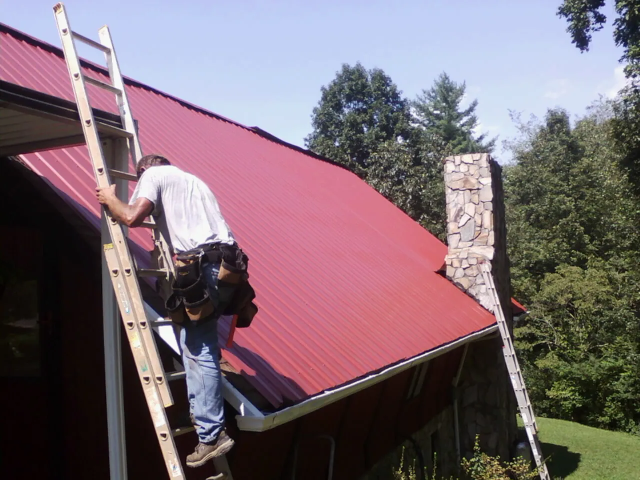 Roofer on a ladder beside a red metal roof and stone chimney — Soffit & Fascia Repair by Roofing Brothers in Locust